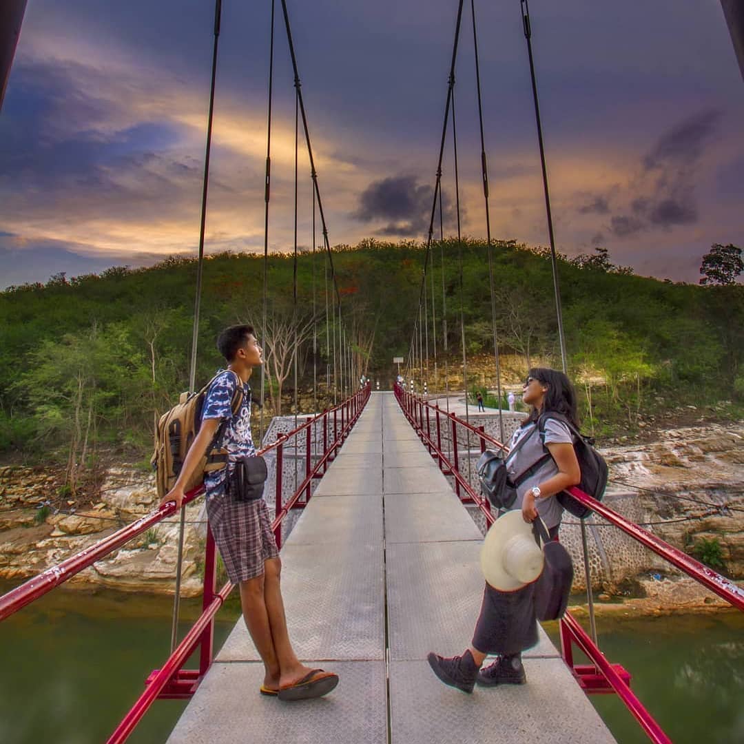 Two visitors standing on a visually stunning suspension bridge in Wanagama Forest during sunset, surrounded by lush greenery and rocky terrain.