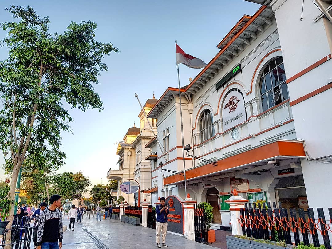 Yogyakarta Post Office building with the Indonesian flag flying above, located near Kilometer Zero. People are walking along the well-maintained pedestrian area beside the historic office.