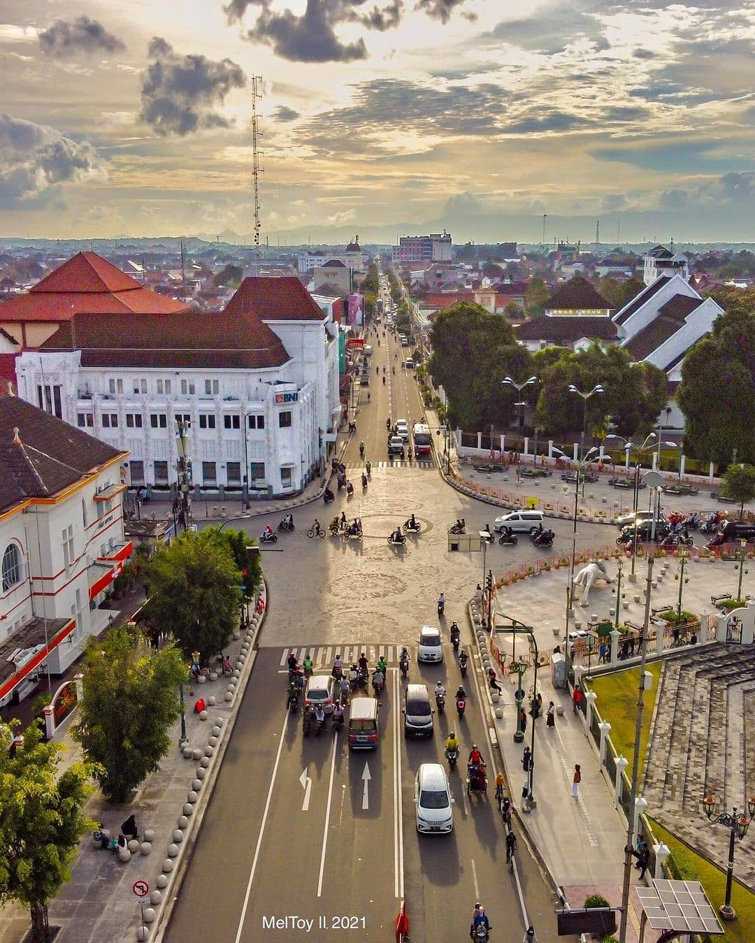 An aerial view of the Yogyakarta Kilometer Zero intersection with cars, motorbikes, and pedestrians. The BNI building and Yogyakarta Post Office can be seen, as well as a long street stretching into the distance under a cloudy sunset sky.