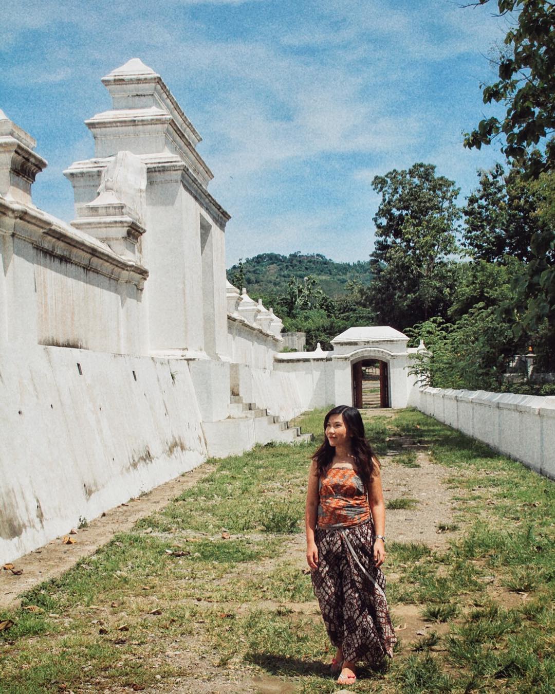 A woman in traditional Javanese attire walks along the white walls of Imogiri Royal Cemetery, under a bright blue sky.