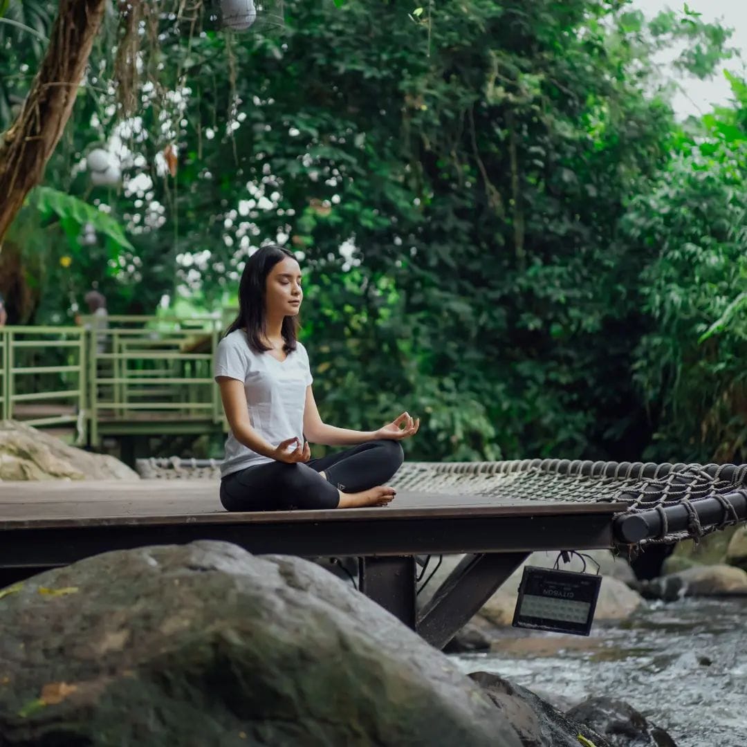 A woman meditating in a peaceful yoga pose on the Secret River Deck at Obelix Village, surrounded by lush greenery and the gentle sound of the flowing river below.