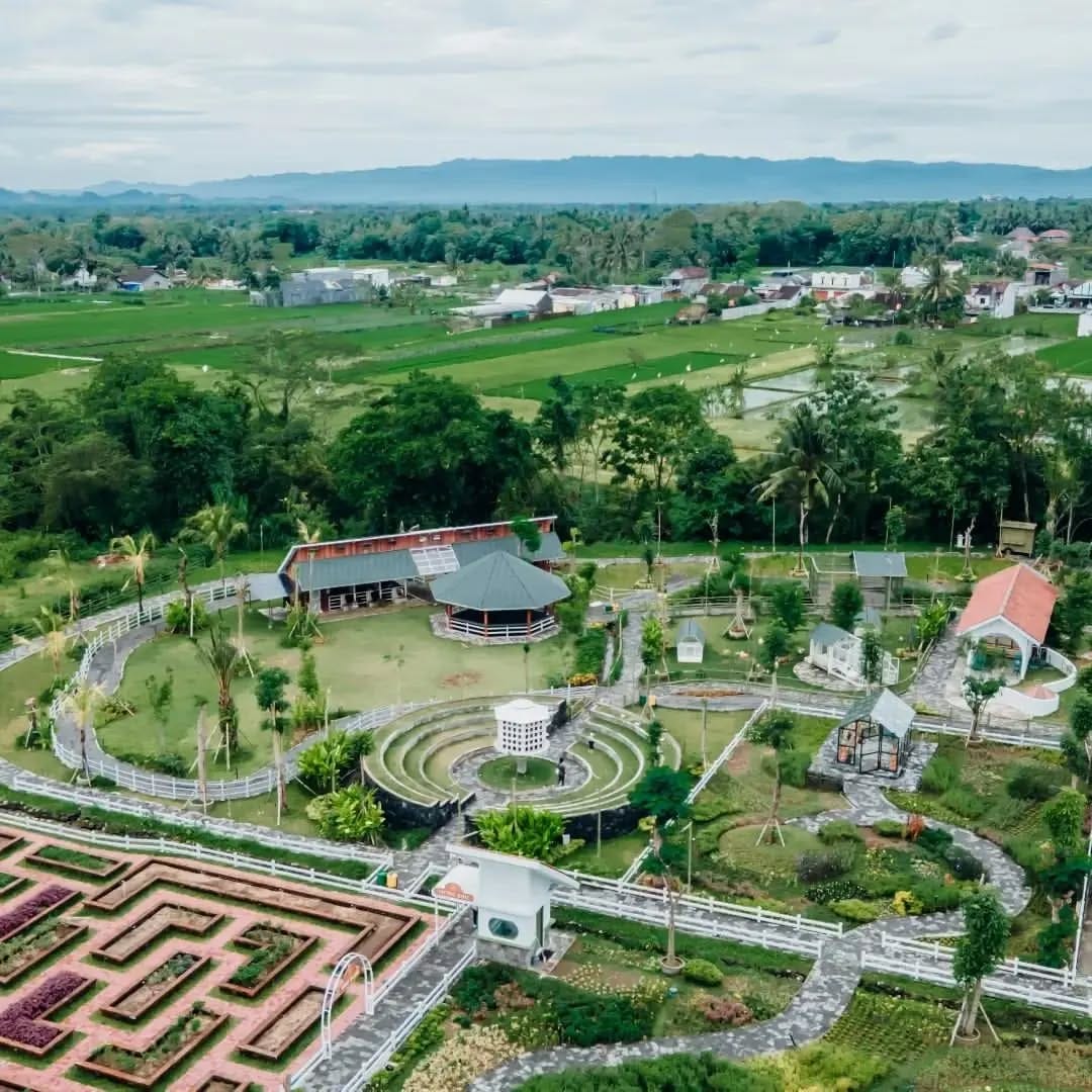 A vibrant aerial view of Obelix Village in Yogyakarta, featuring a beautifully designed maze garden, lush greenery, walking paths, and traditional-style buildings surrounded by rice fields.