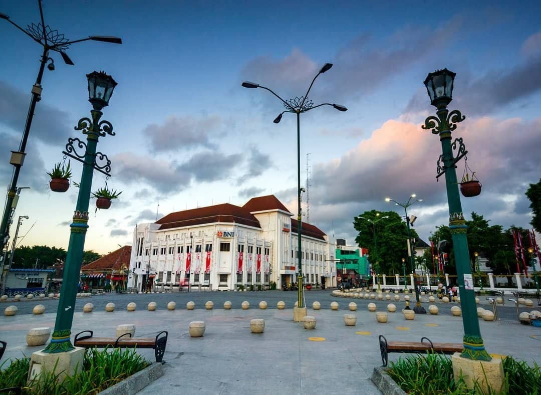 The BNI building at Yogyakarta Kilometer Zero during early evening, framed by ornate street lamps and stone bollards. The sky is tinged with hues of sunset, creating a peaceful scene at the landmark.