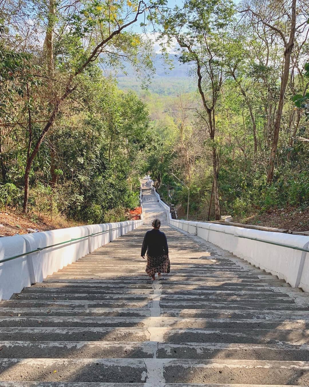 A person in traditional Javanese clothing walking up a long, steep staircase surrounded by trees at Imogiri Royal Cemetery.