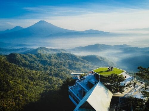 A breathtaking view from Tumpeng Menoreh in Kulon Progo showcasing a hilltop viewing platform with lush green forests below and majestic mountains, including Mount Merapi, in the background under a clear blue sky.