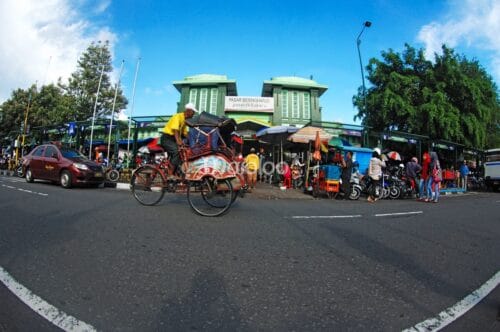 A bustling street in front of the west entrance of Beringharjo Market, with a man riding a traditional becak (pedicab) and people shopping at roadside stalls under colorful umbrellas.