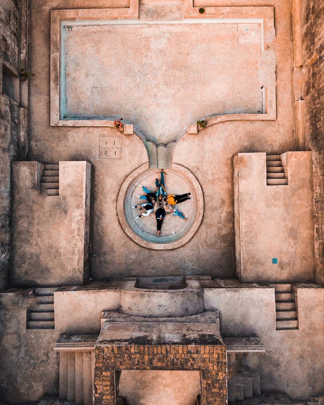 A group of friends lies in a circle at the center of the round stone pond at Warungboto Site, viewed from above, surrounded by ancient architectural structures.