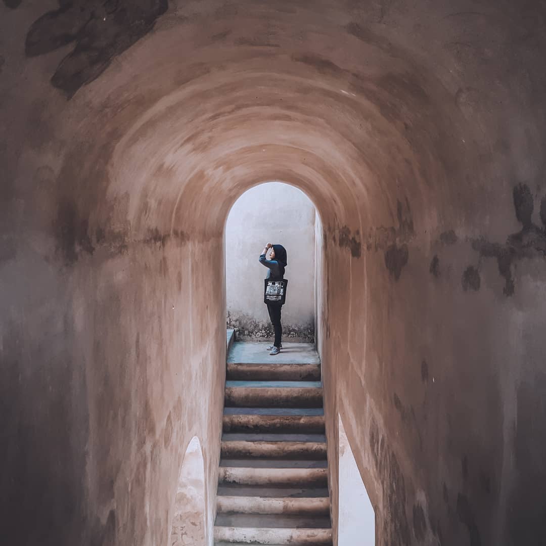 A visitor stands inside the arched corridor of Warungboto Site, Yogyakarta, with stairs leading to an intriguing passageway framed by ancient stone walls.