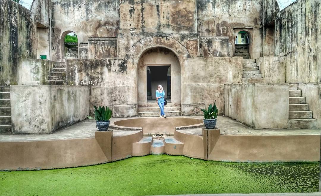 A visitor stands in front of the iconic pool and grand archway at Warungboto Site in Yogyakarta, surrounded by ancient stone structures and moss-covered water.