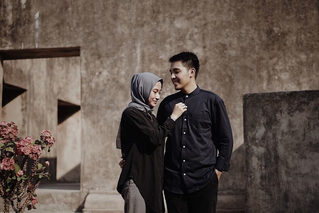 A couple poses for a pre-wedding photo at Warungboto Site in Yogyakarta, standing in front of ancient stone walls and a blooming plant.