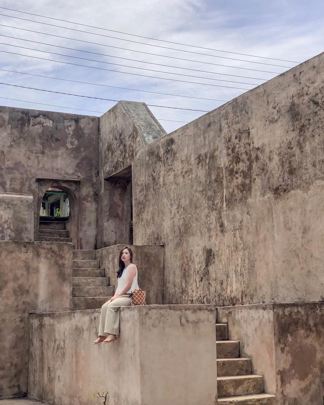 A woman sits on a stone ledge at Warungboto Site in Yogyakarta, surrounded by ancient brick walls and staircases under a cloudy sky.