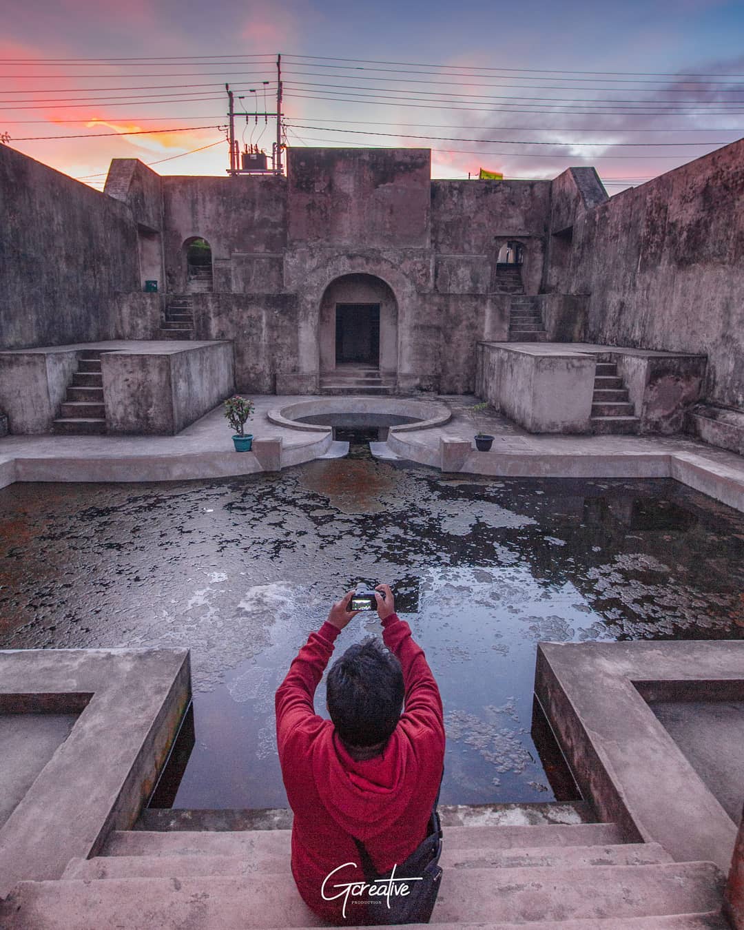 A photographer in a red hoodie captures the stunning Warungboto Site in Yogyakarta during sunset, with the reflection of the sky on the central pool.